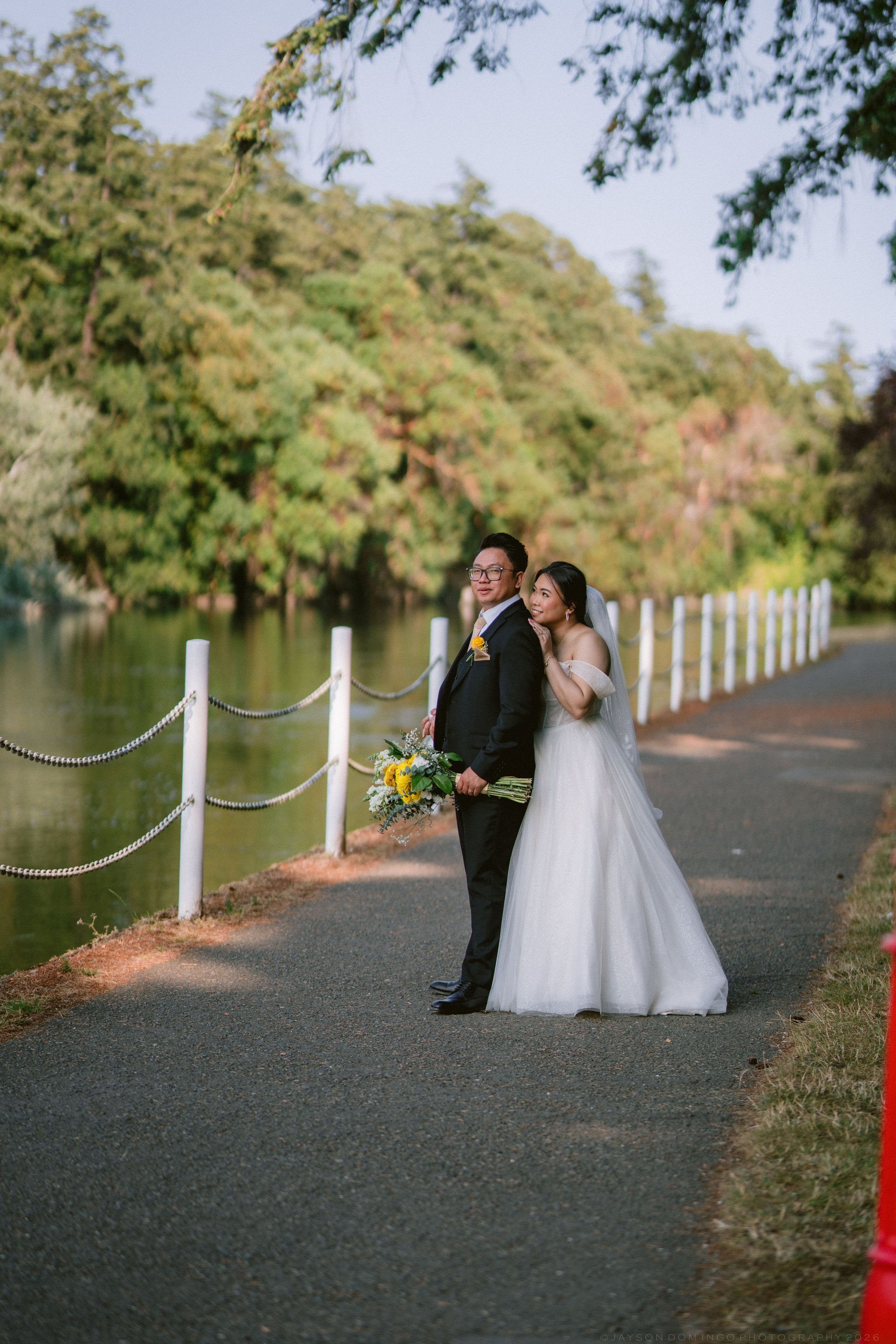 Bride and groom pose on lakeside path surrounded by lush green trees on their wedding day.