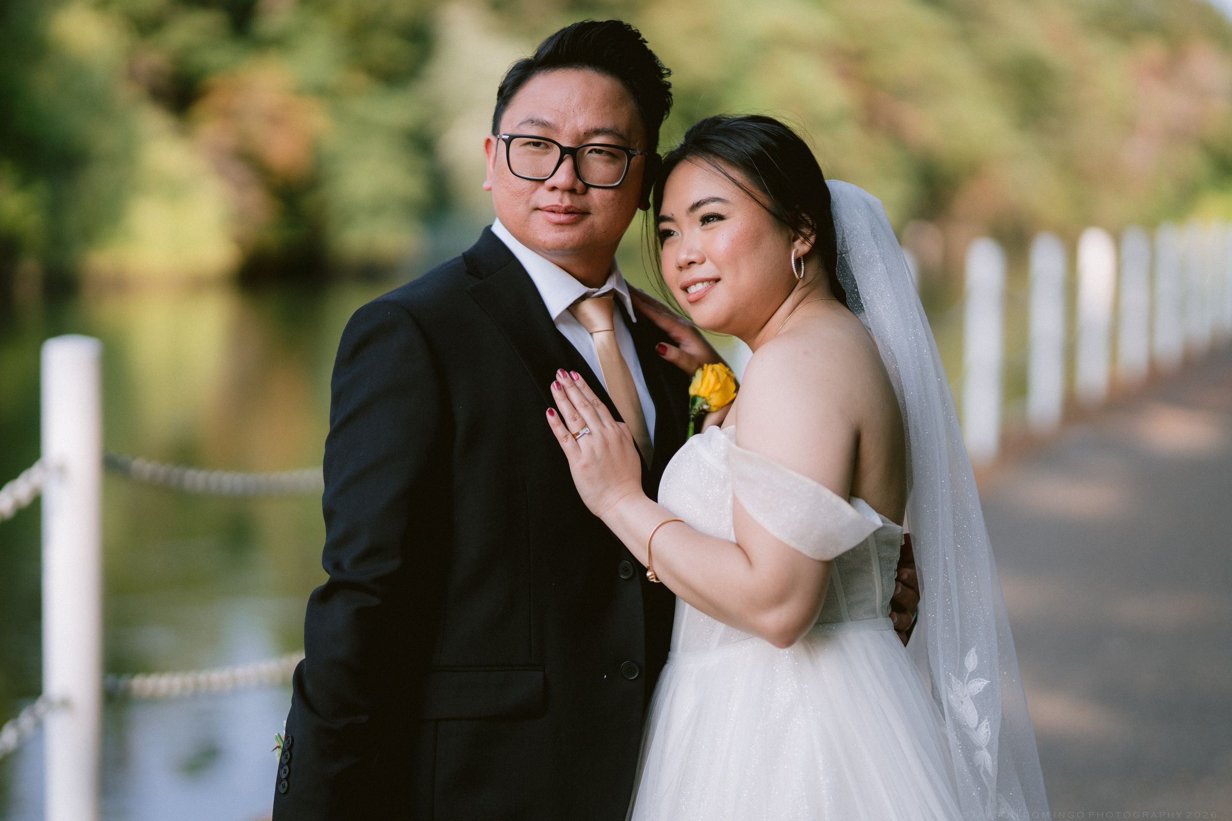Bride and groom share a romantic pose on a lakeside walkway, bride in white gown with yellow flower.