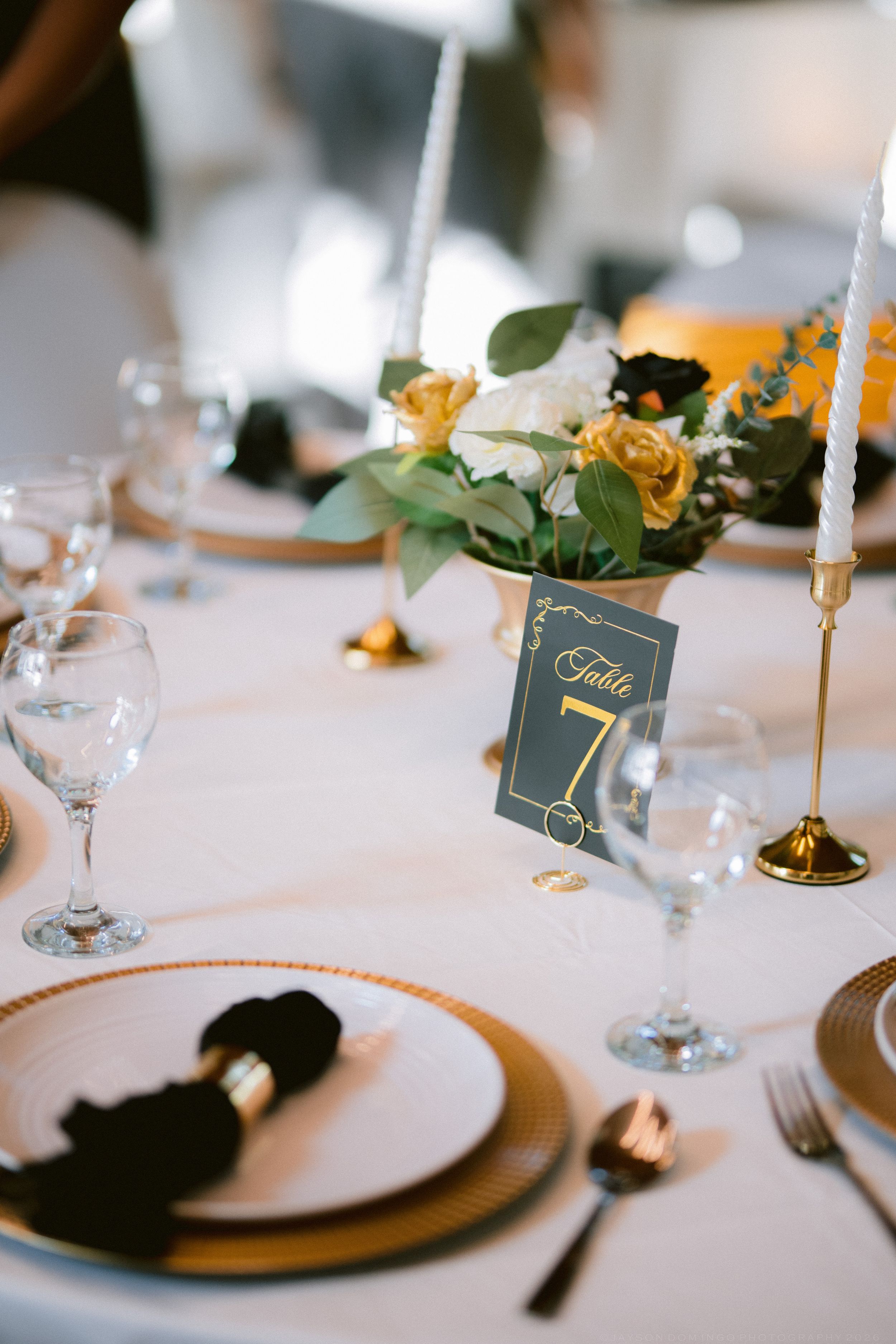 Elegant wedding table setting with gold chargers, black napkins, yellow roses, and Table 7 card.