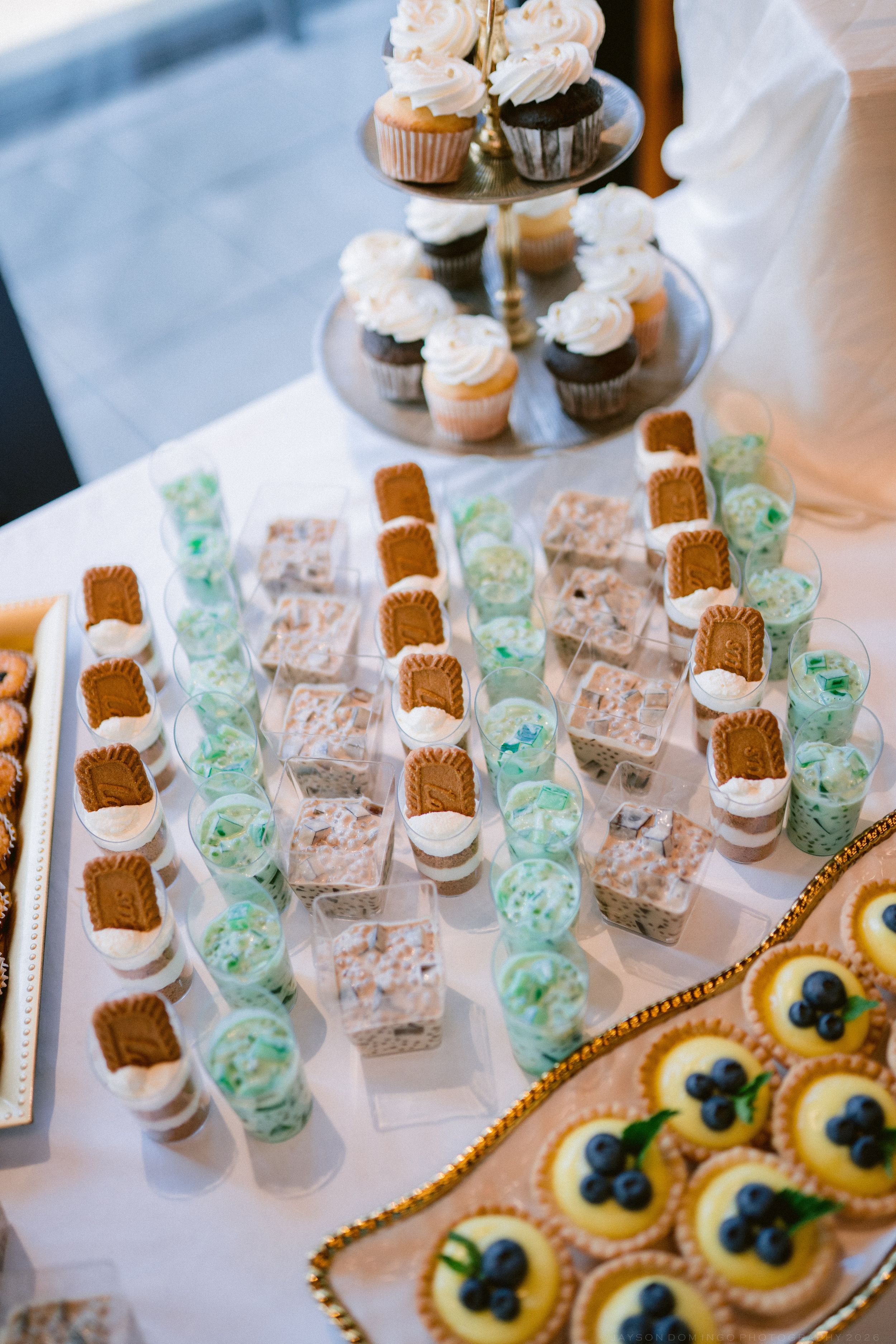 Elegant wedding dessert table featuring cupcakes, cookie cups, mint parfaits, and blueberry tarts on white linen.