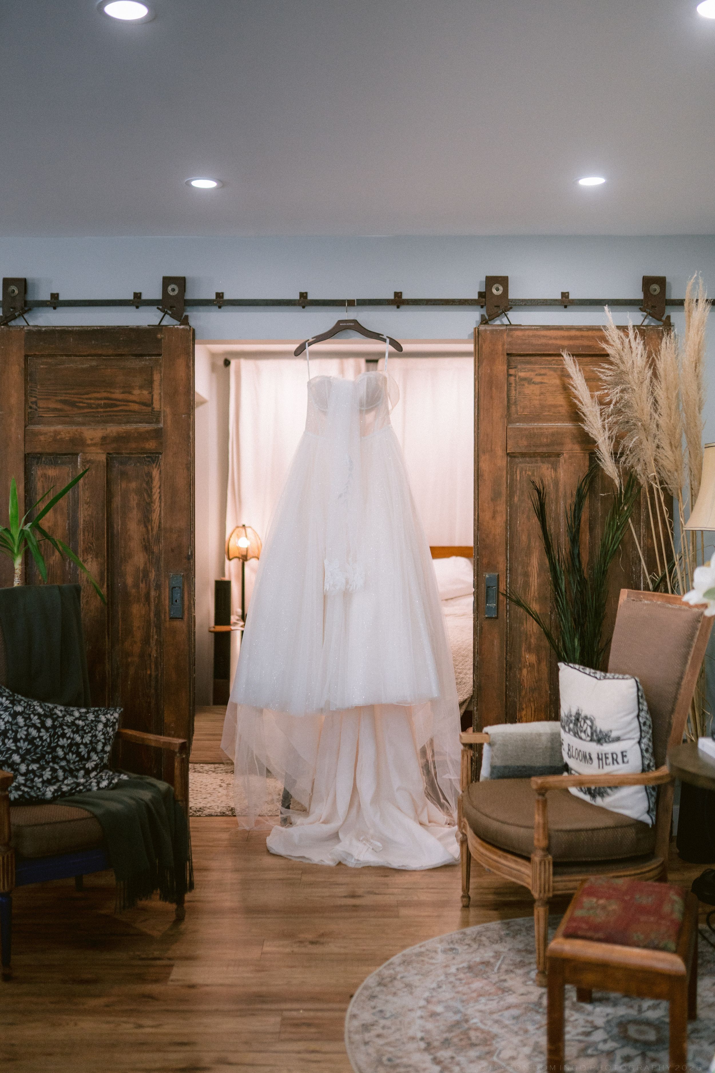 White wedding dress hanging on rustic barn doors in a cozy bridal suite with wooden floors.