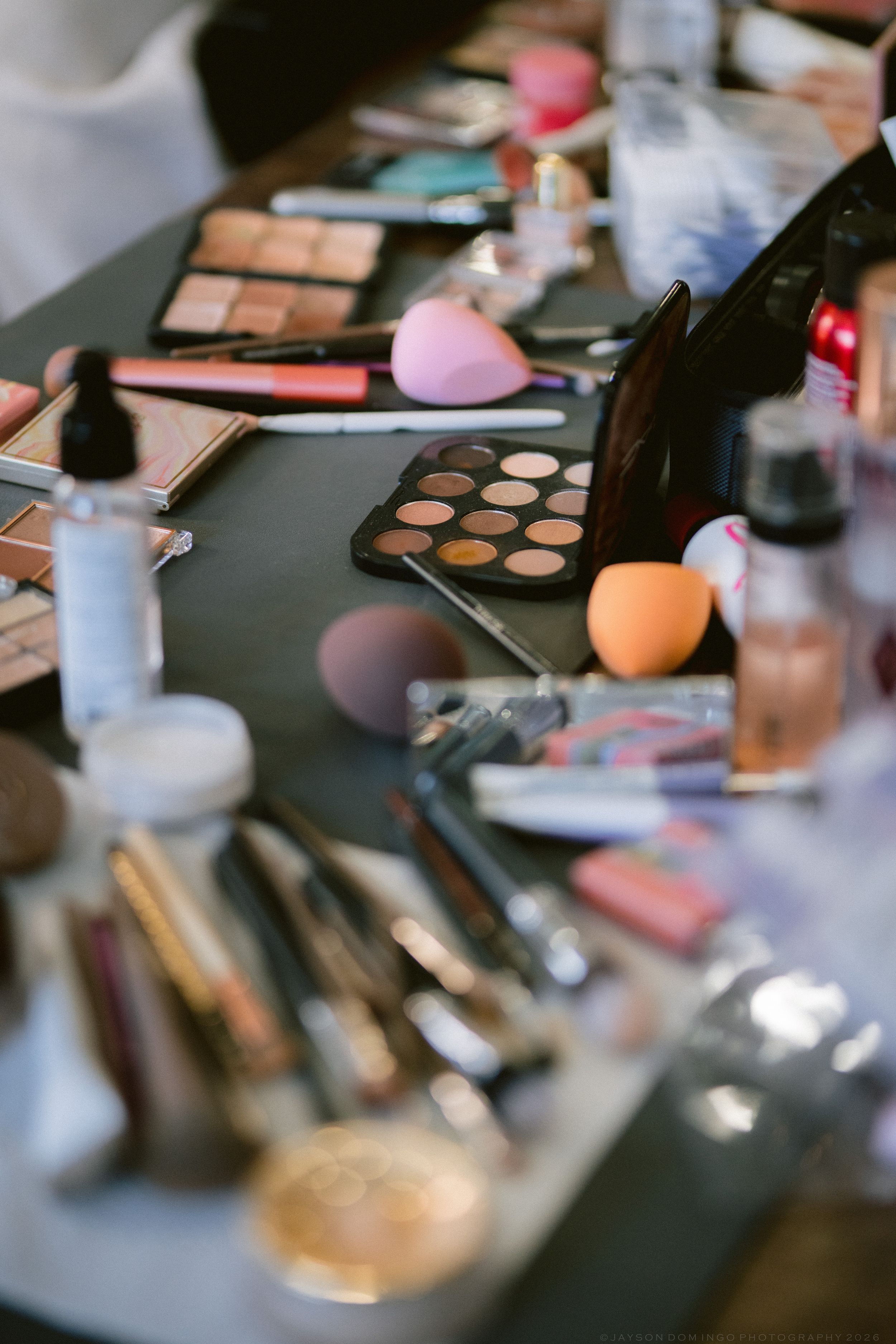Cluttered makeup table with eyeshadow palettes, brushes, beauty blenders, and cosmetics for wedding day preparation.