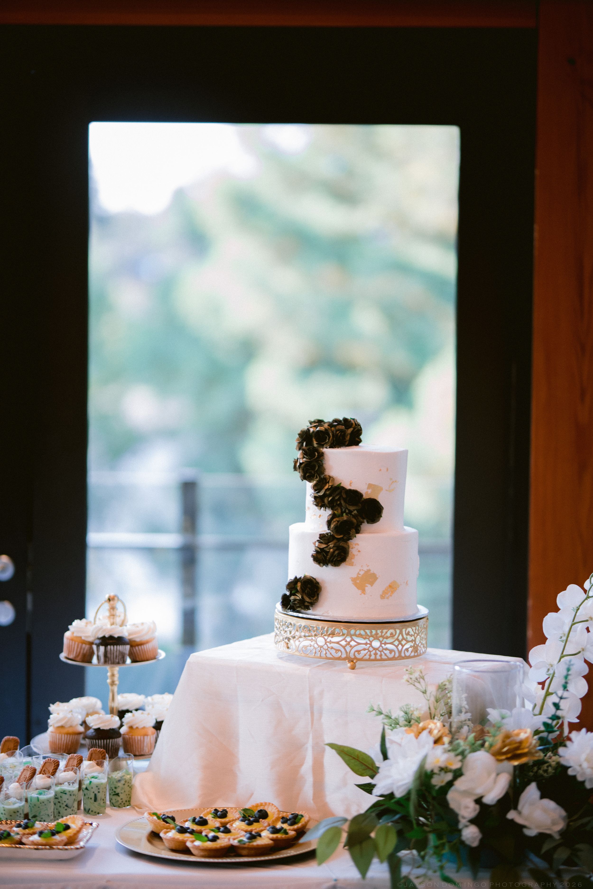 Three-tier white wedding cake with black floral accents and gold details displayed on ornate stand with florals.