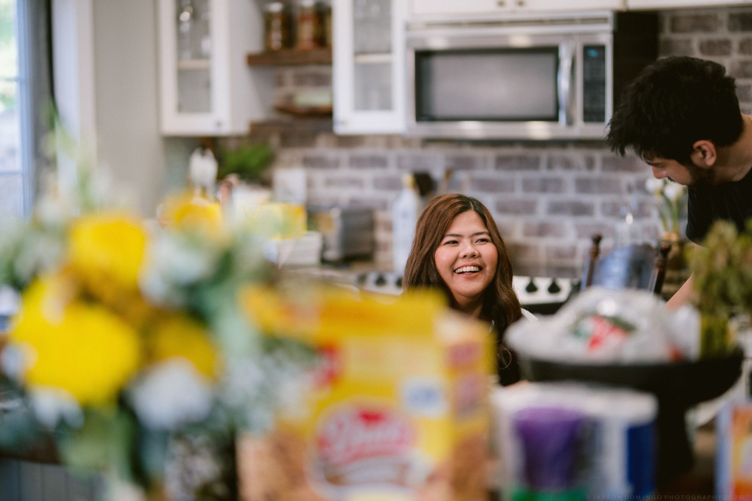 A smiling woman laughs in a modern kitchen with white cabinets, a microwave, and brick backsplash.