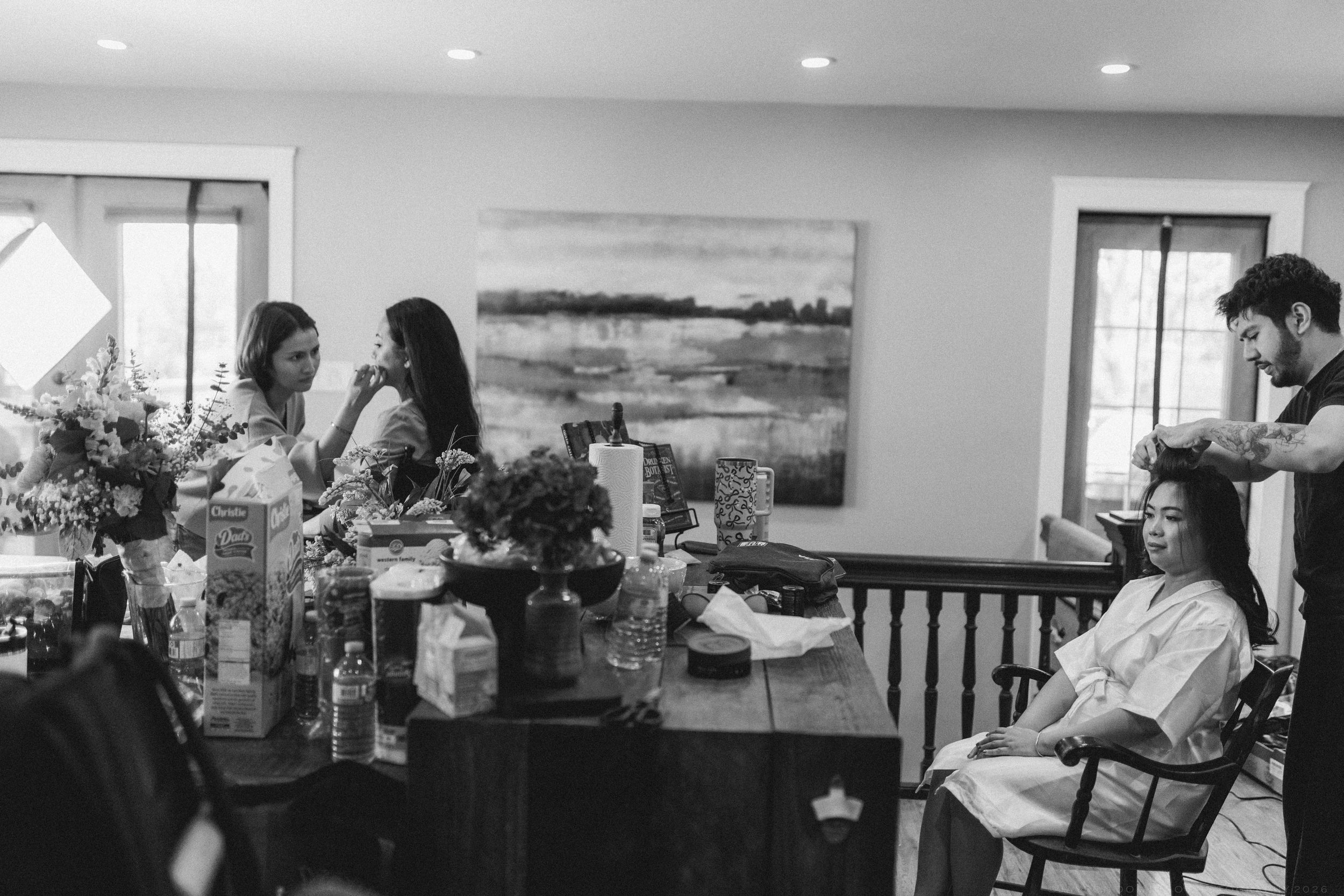 Bride getting hair done by stylist while bridesmaids chat at table with floral arrangements in cozy home setting.