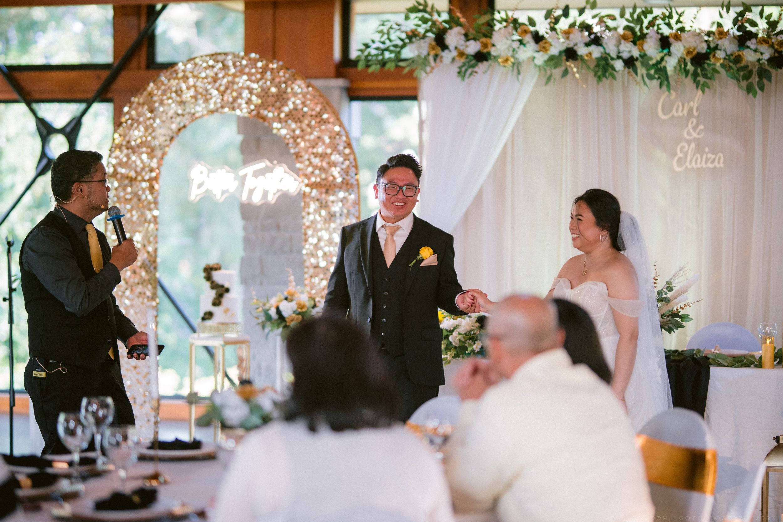 Elegant wedding ceremony with groom in black suit, bride in white off-shoulder gown, and officiant at floral arch venue.