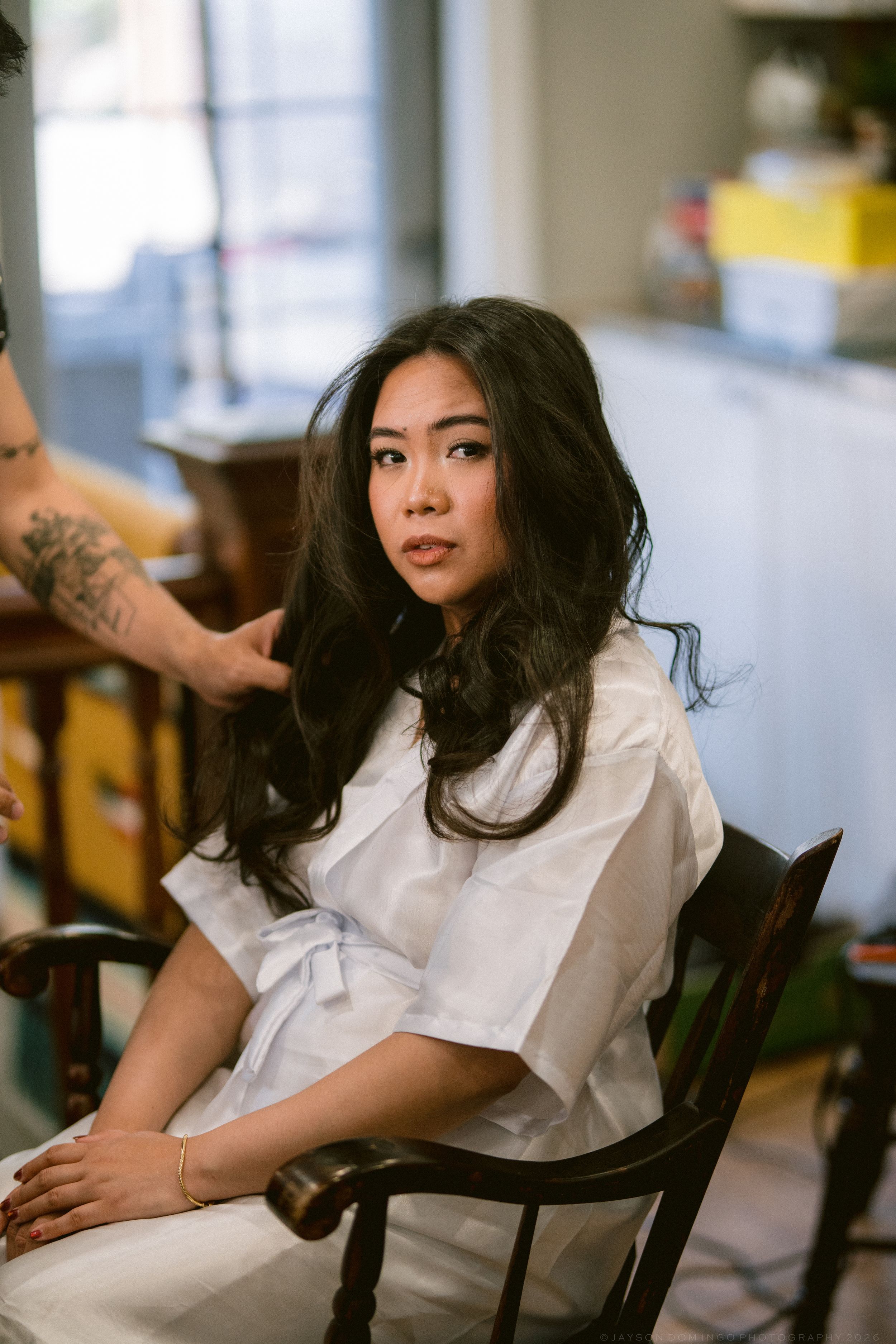 Asian bride with long wavy hair getting ready in a white robe while a stylist works on her hair.
