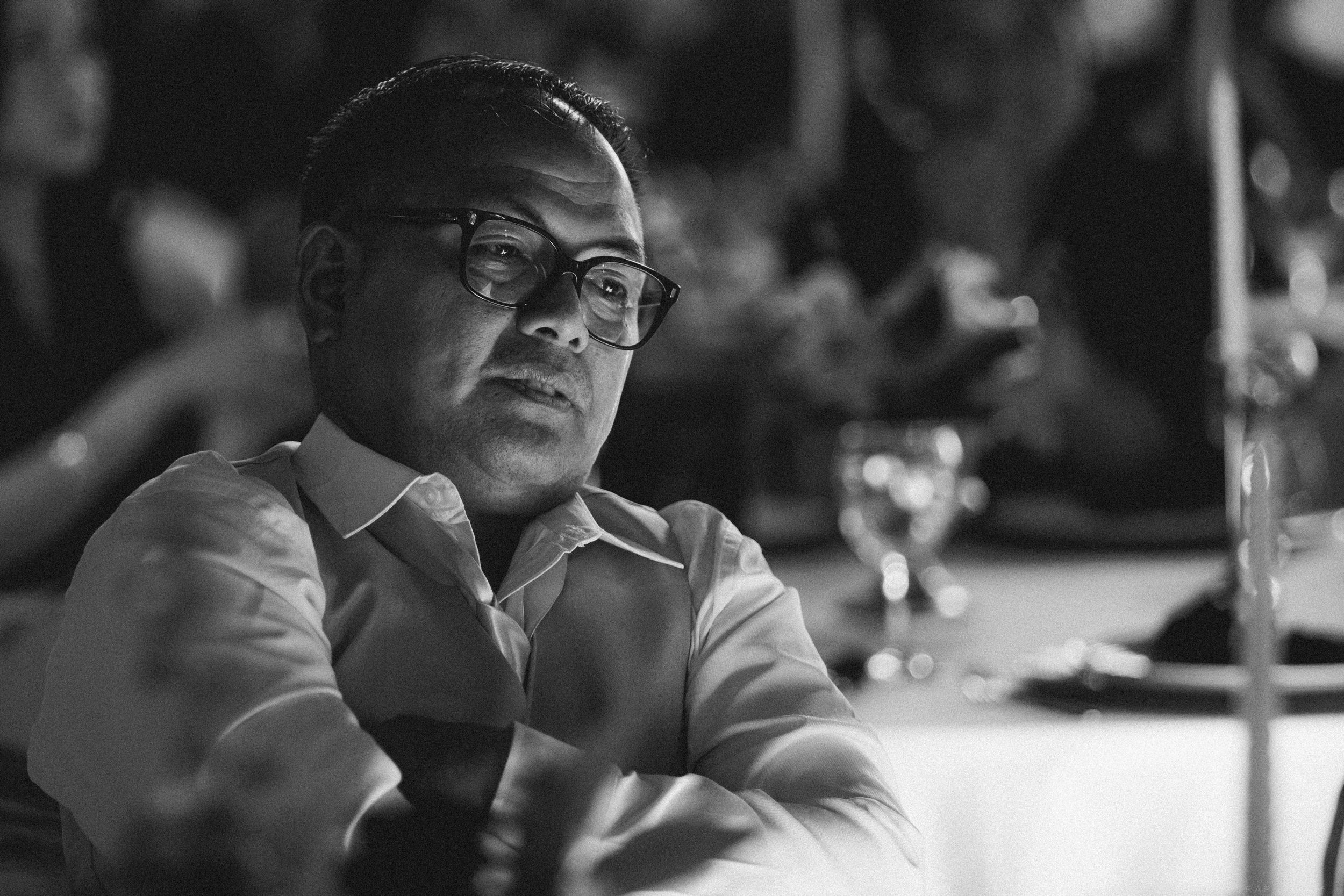 Black and white close-up portrait of man wearing glasses seated at wedding reception table.