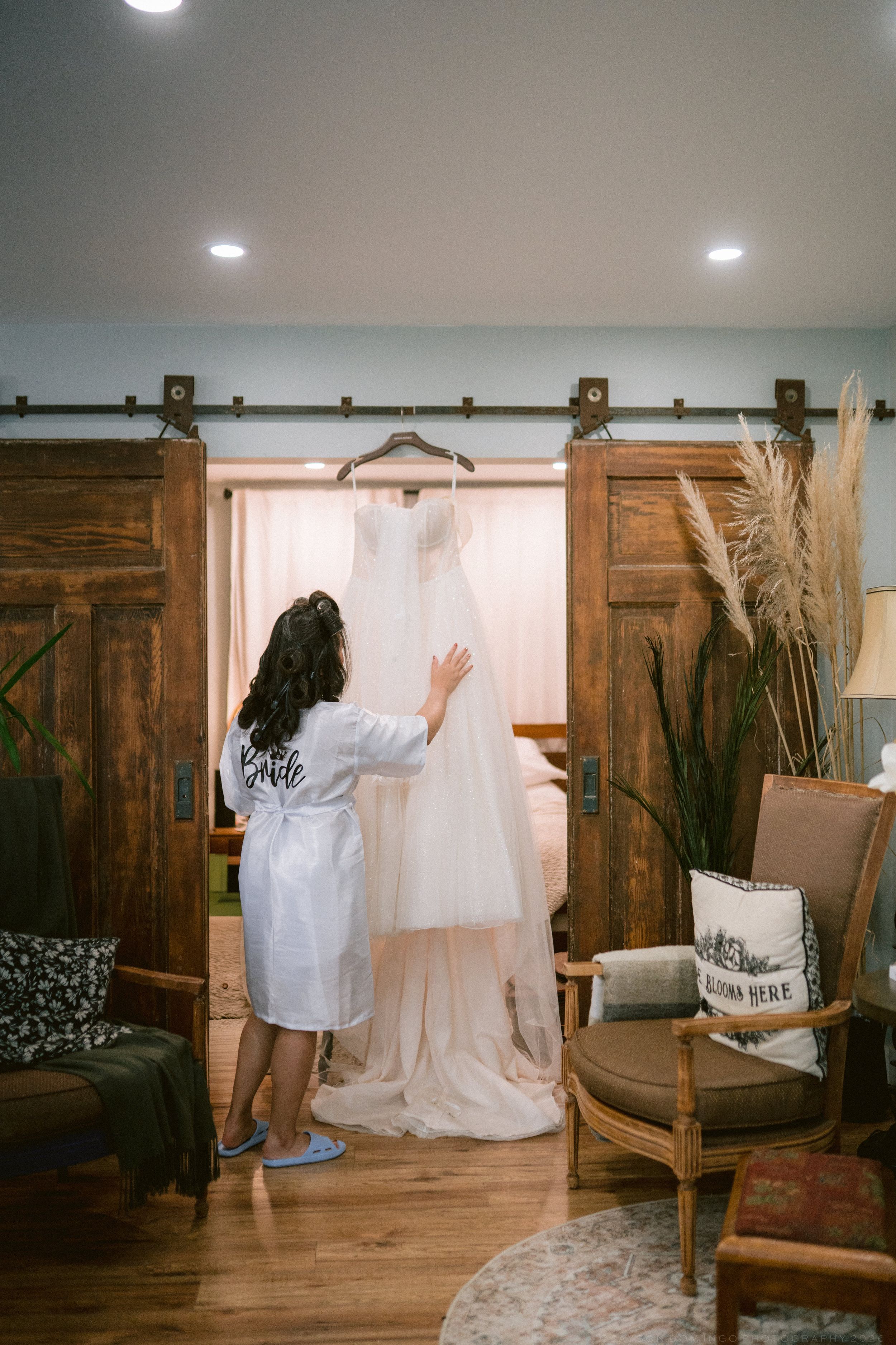 Bride in white robe touching her wedding gown hanging on rustic barn doors before ceremony.