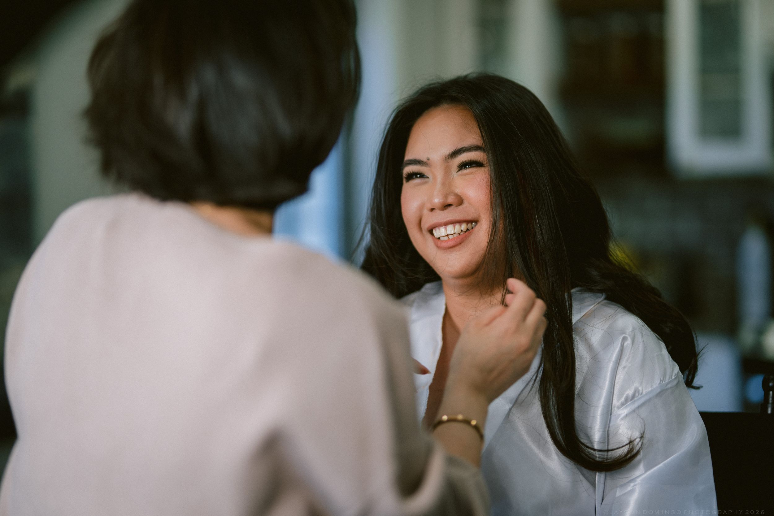 Smiling Asian woman with long black hair laughing during a conversation with another person in a bright indoor setting.