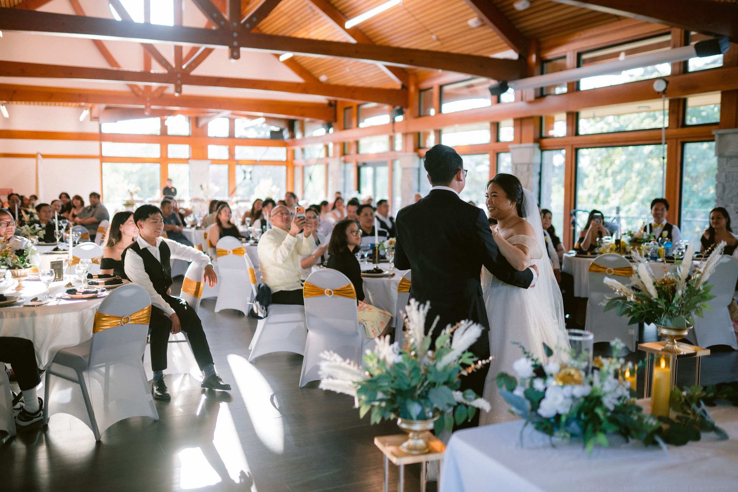 Wedding reception in wooden venue with guests seated at white-clothed tables while bride and groom share first dance.