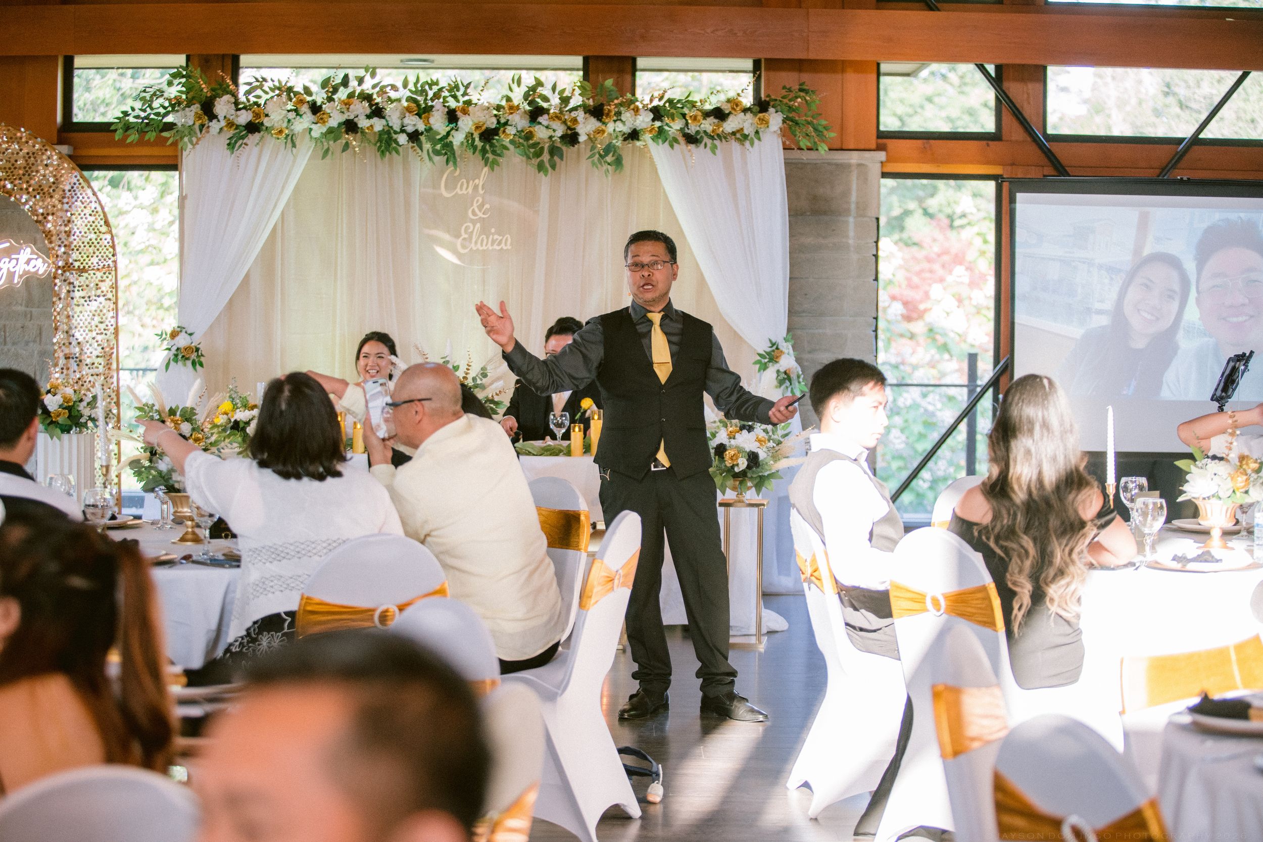 Wedding MC in black suit gesturing to guests in a decorated reception venue with floral arch.