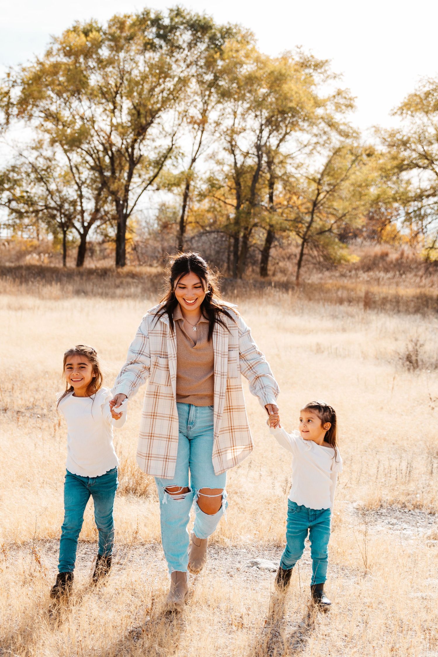 A smiling woman in plaid jacket and jeans walks hand-in-hand with two young girls in a golden autumn field.