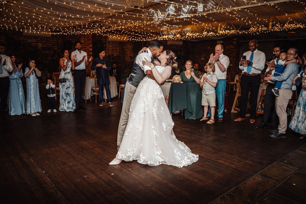 Bride and groom share first dance surrounded by applauding guests in a warmly lit rustic wedding venue.