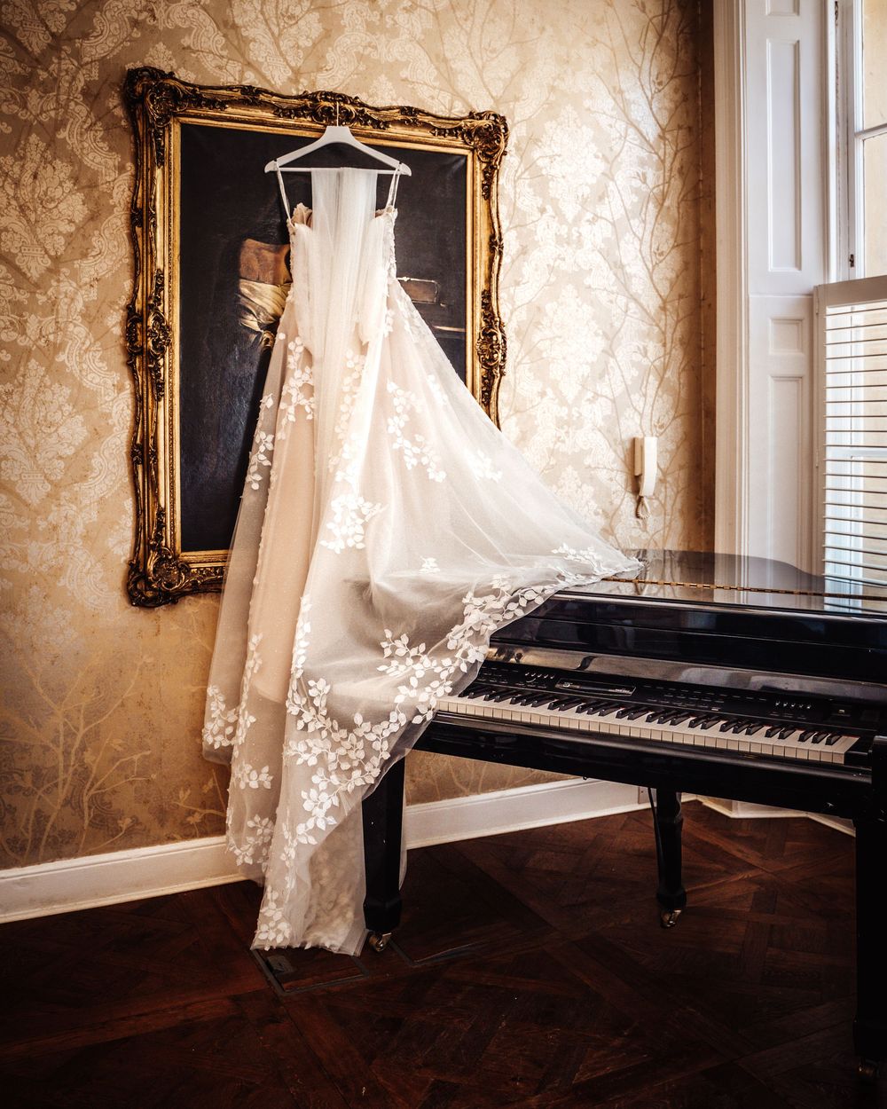 Elegant floral lace wedding dress hanging near a grand piano against ornate gold-framed artwork.