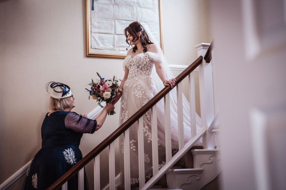 Bride in lace wedding gown holding bouquet on staircase while woman in black adjusts her dress.