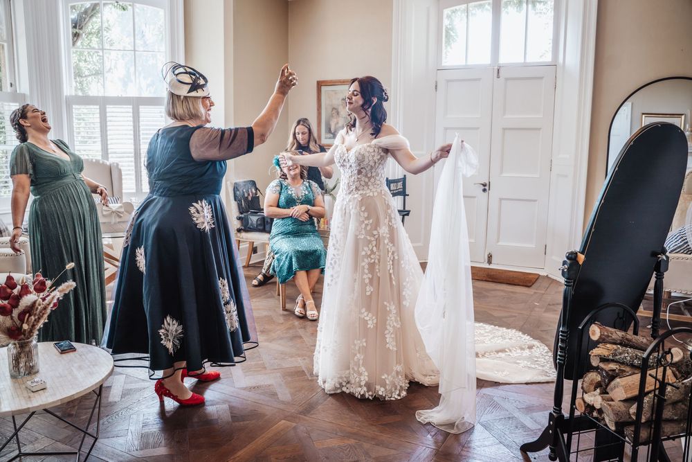 Bride in lace gown gets veil adjusted by guests in elegant sunlit room.