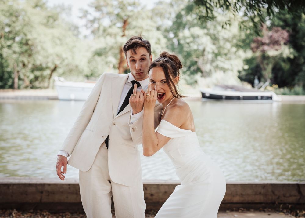 Joyful bride and groom posing playfully by a scenic lakeside, groom in cream suit, bride in off-shoulder gown.