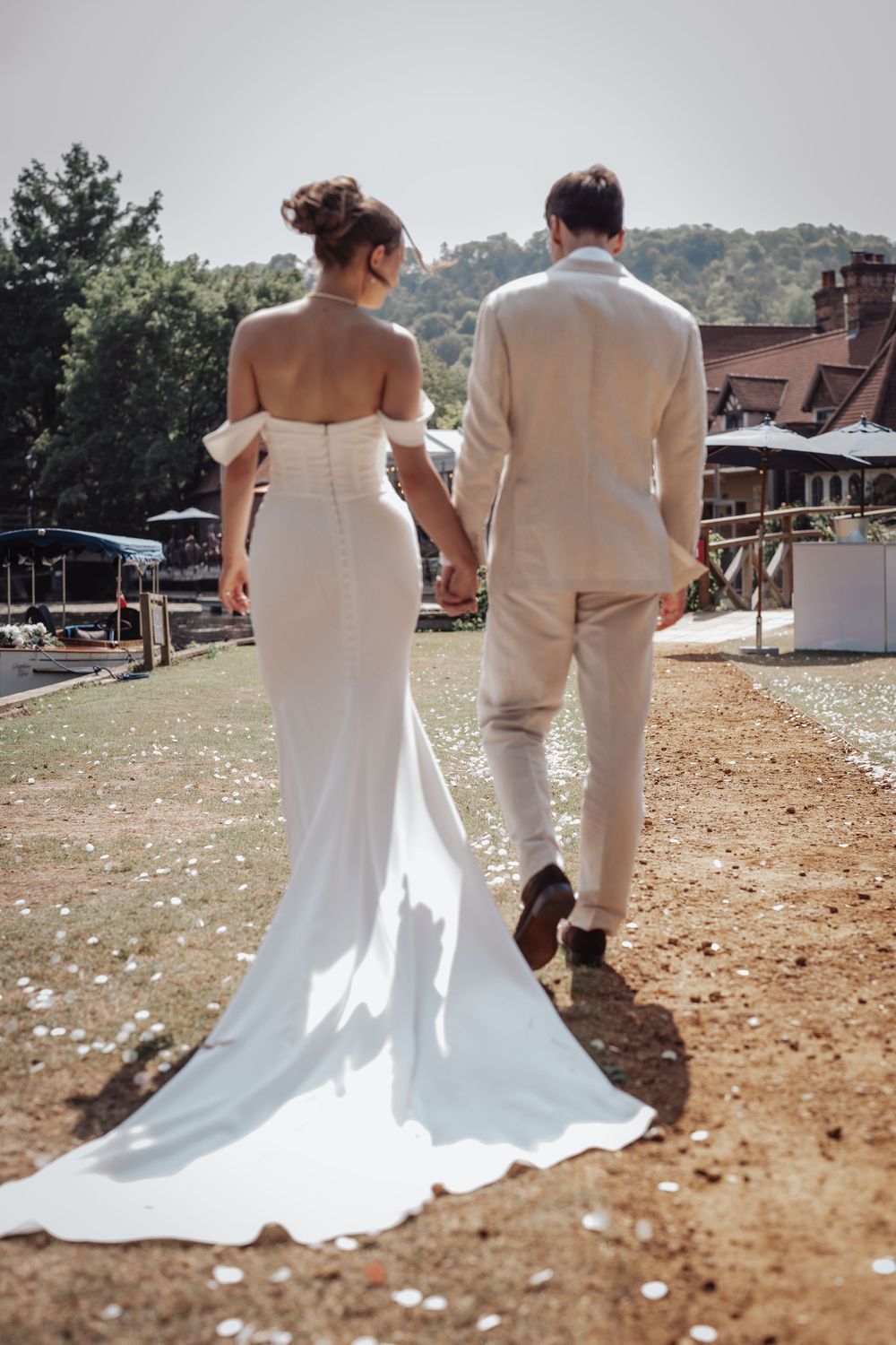 Bride and groom walking hand in hand outdoors in sunlight, viewed from behind on wedding day.