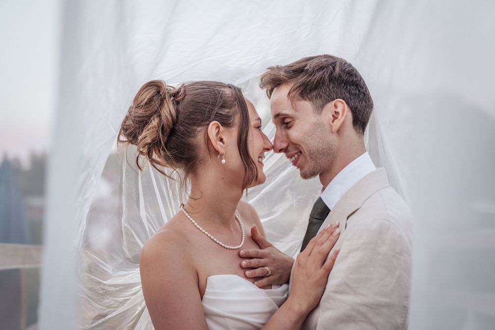 Smiling newlyweds share intimate moment wrapped in flowing white veil outdoors.