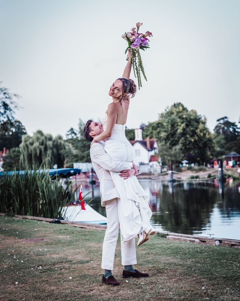 Groom lifts bride holding colorful bouquet high in joyful wedding photo by scenic lakeside.