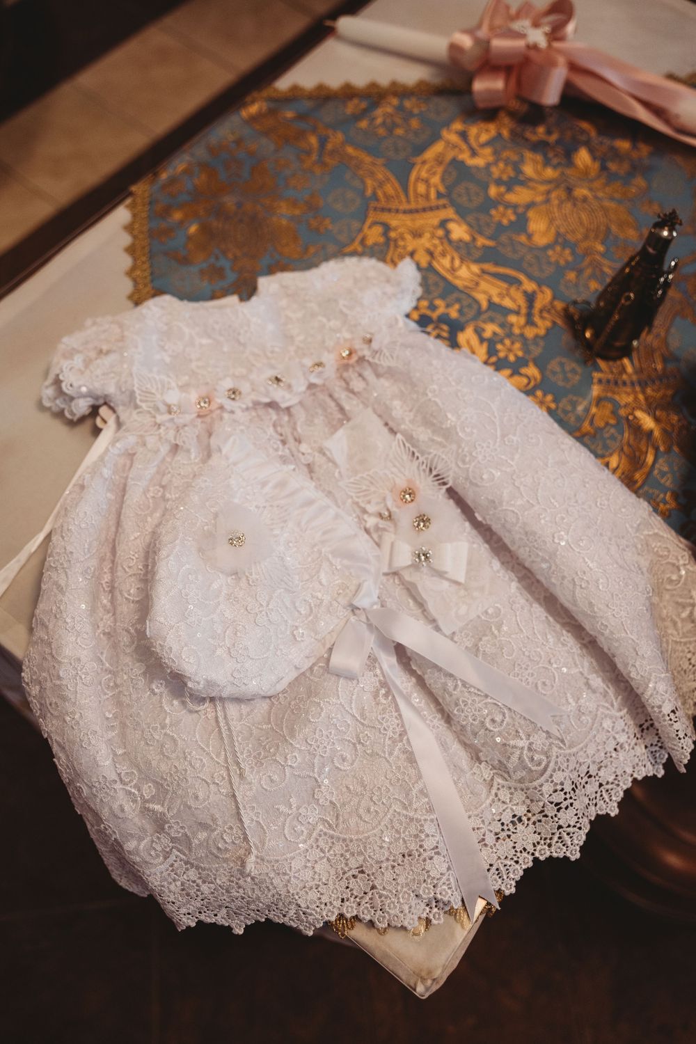 Delicate white lace christening gown and bonnet with satin ribbons displayed on ornate gold and blue fabric.