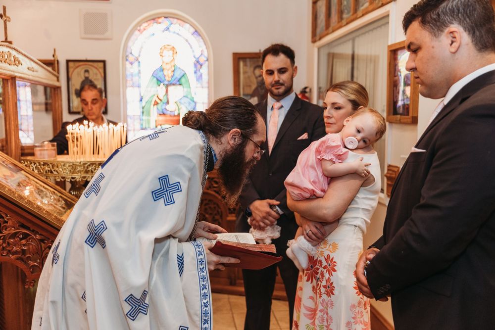 Orthodox priest blessing a baby during a christening ceremony in a church with stained glass windows.
