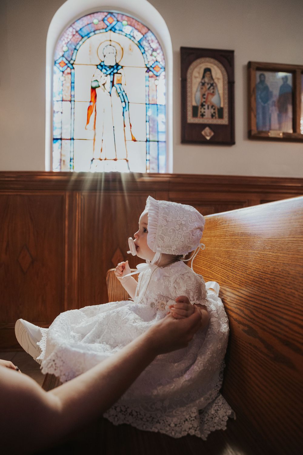 Baby in white lace christening gown and bonnet sitting on church pew near stained glass window.