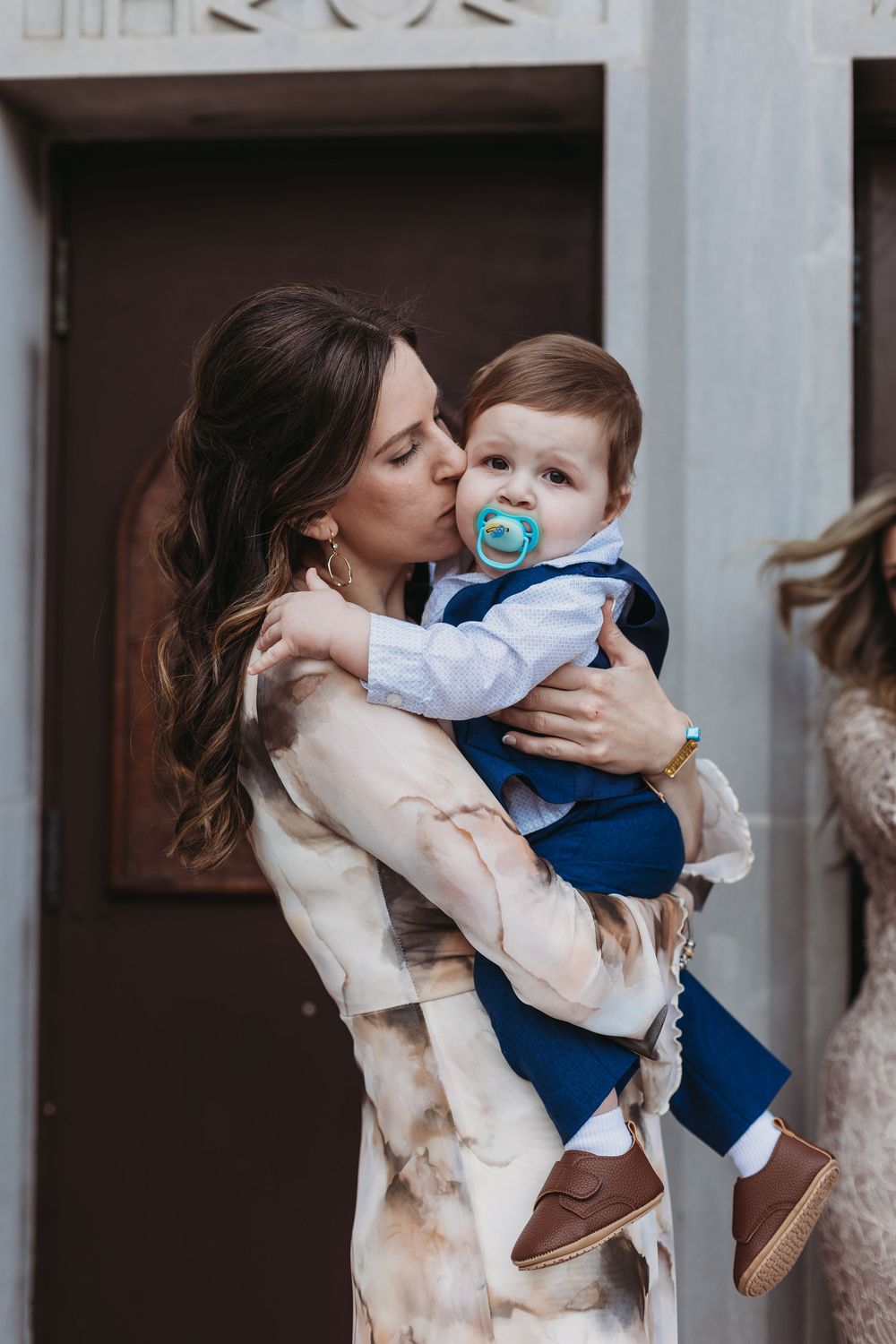 A mother tenderly kisses her baby boy dressed in a blue suit while holding him outside a church.