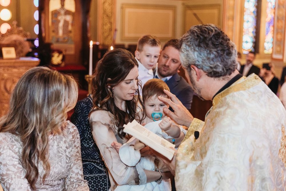 Priest performing a baptism ceremony on an infant held by mother in an ornate Orthodox church.
