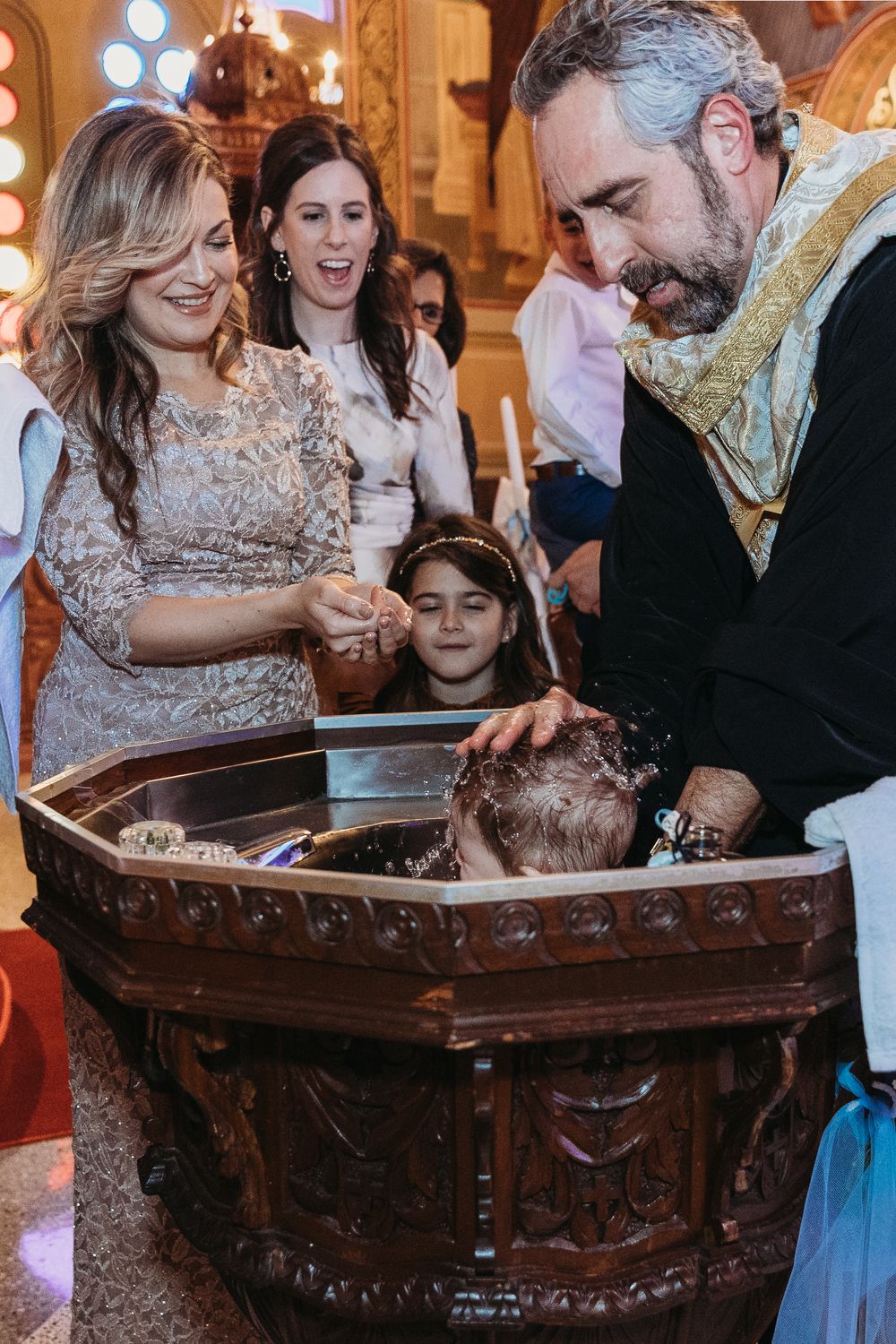 A priest performs an Orthodox Christian baptism ceremony with family gathered around an ornate font.