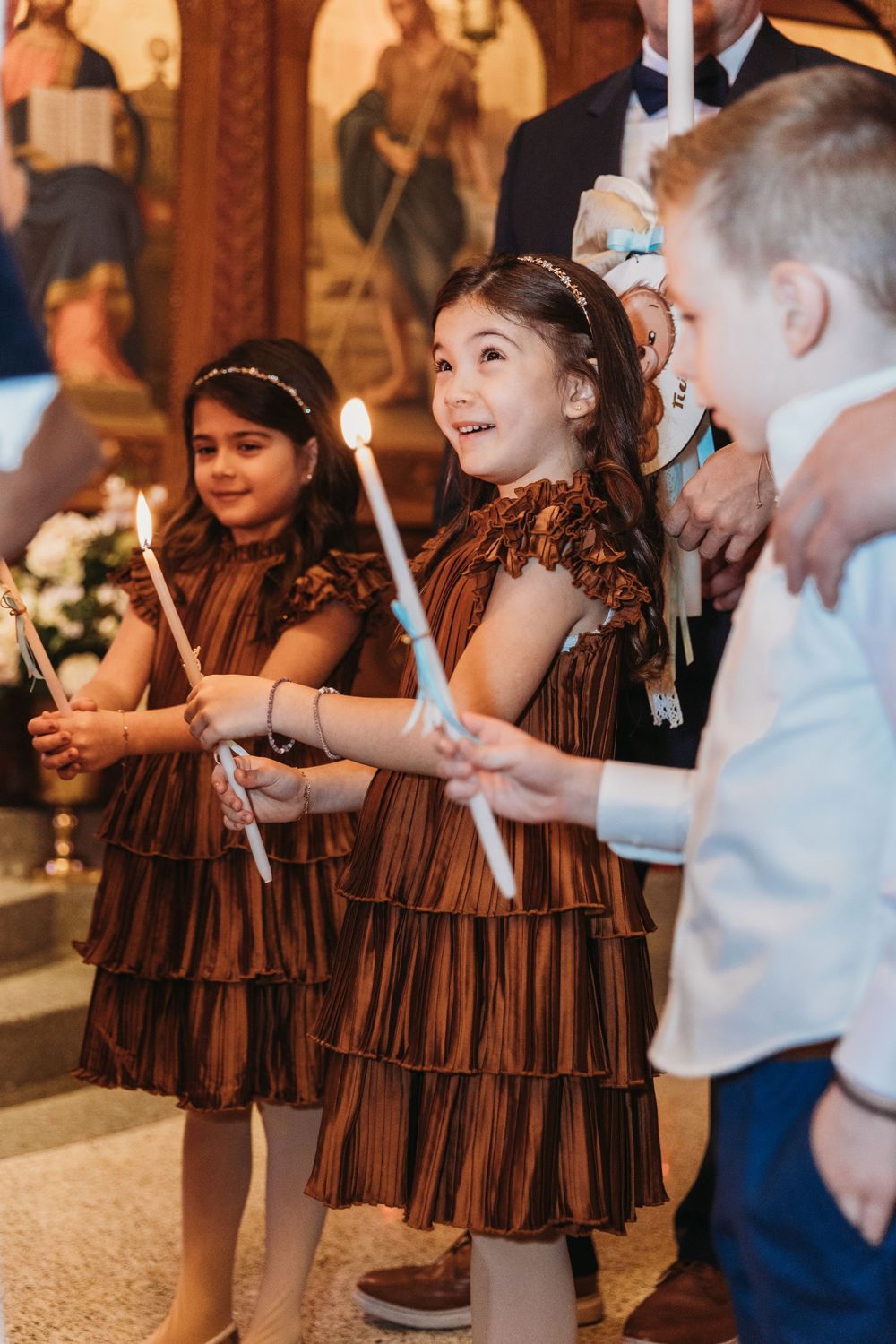 Children in brown dresses holding candles during a church ceremony, smiling joyfully near religious icons.