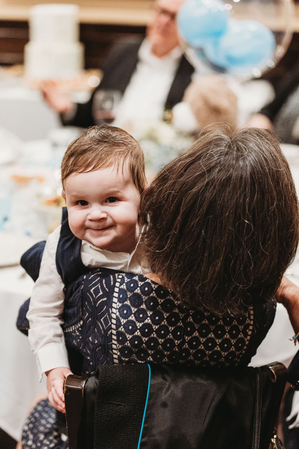 A smiling baby in a white shirt held by an elderly woman at a festive celebration with blue balloons.