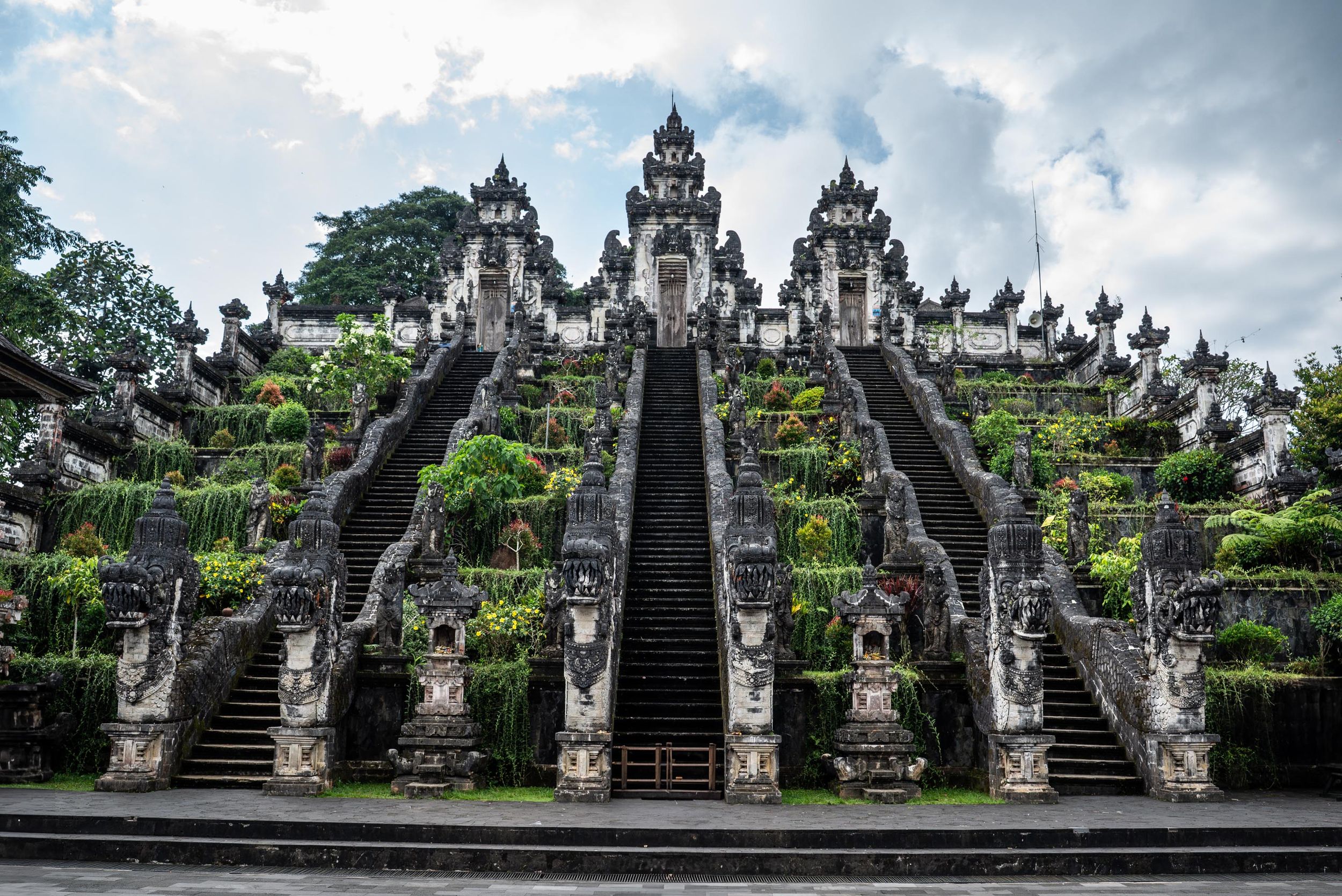Ornate Balinese Hindu temple with intricate stone carvings, tiered staircases, and lush tropical greenery under cloudy skies.