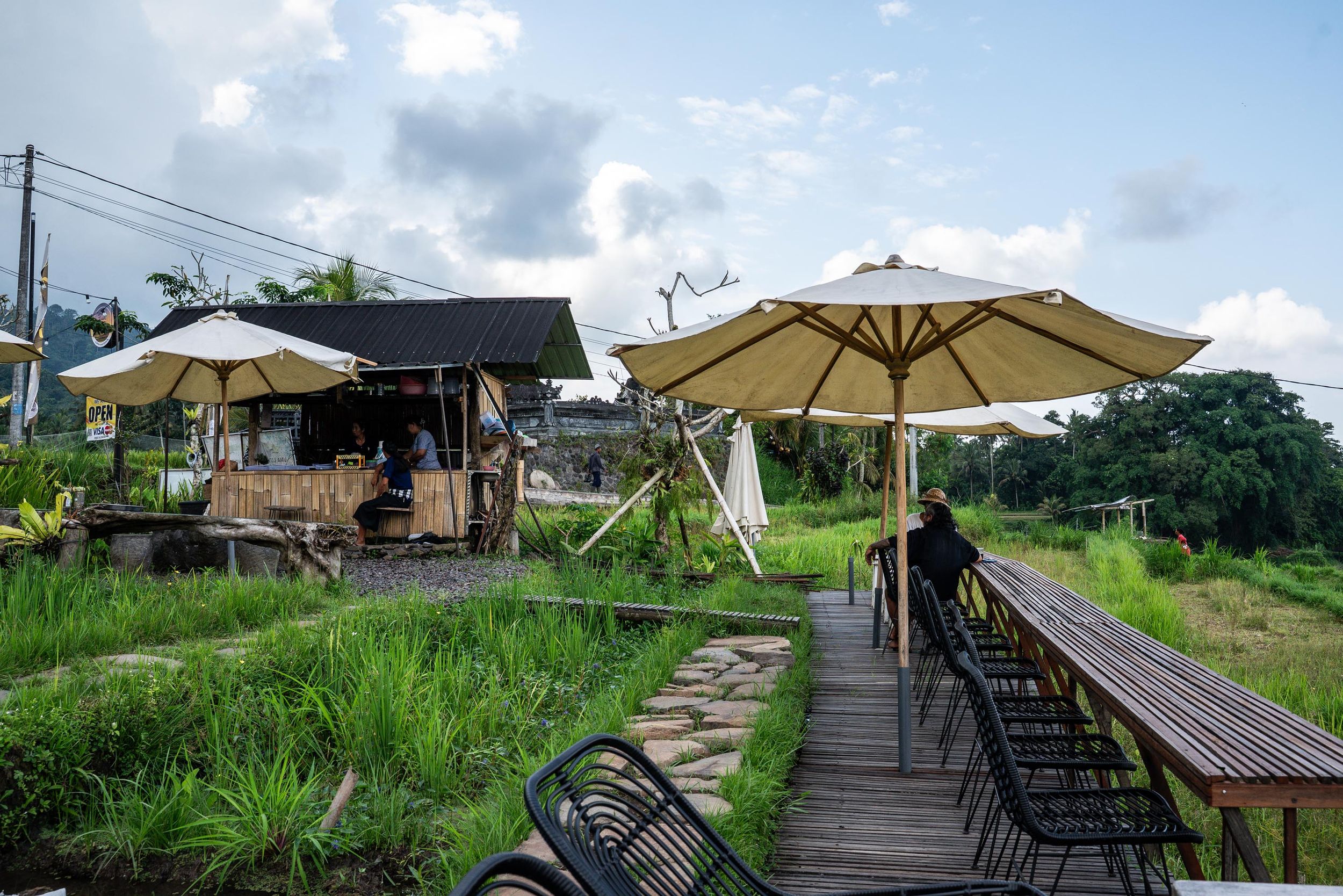 Outdoor cafe with beige umbrellas and wooden deck surrounded by lush greenery under a partly cloudy sky.