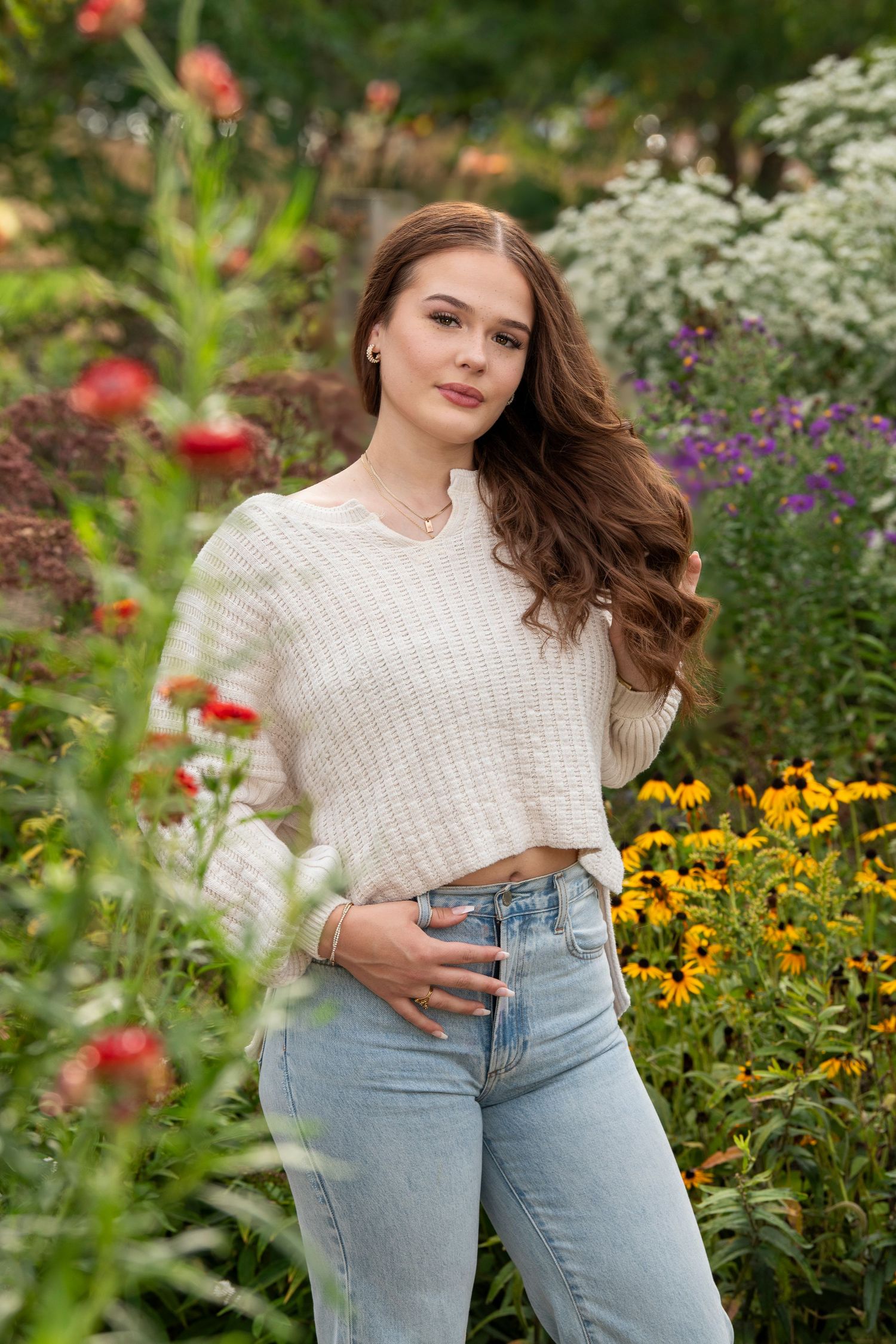 Young woman with long brown hair in white sweater and jeans posing in a colorful garden with flowers.
