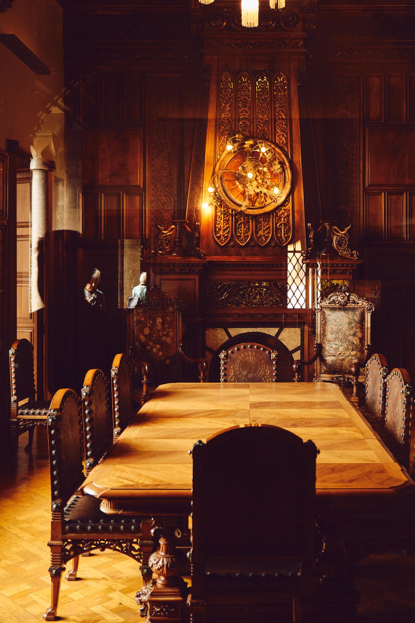 Ornate dark wood dining room with carved fireplace, large round medallion, antique chairs, and parquet floors in a historic mansion.