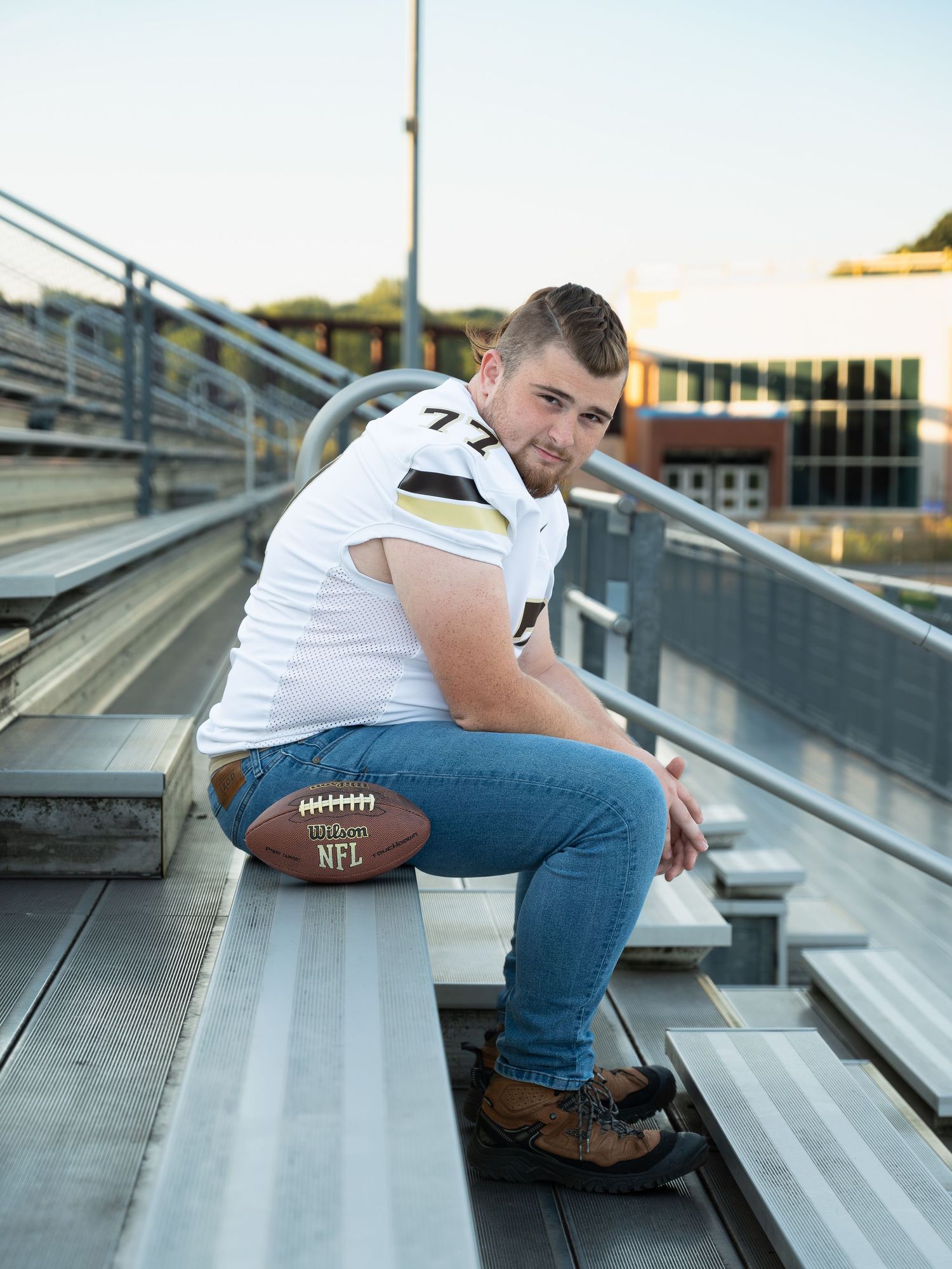 Young football player in football jersey sits on bleachers with football, wearing jeans and boots at stadium.