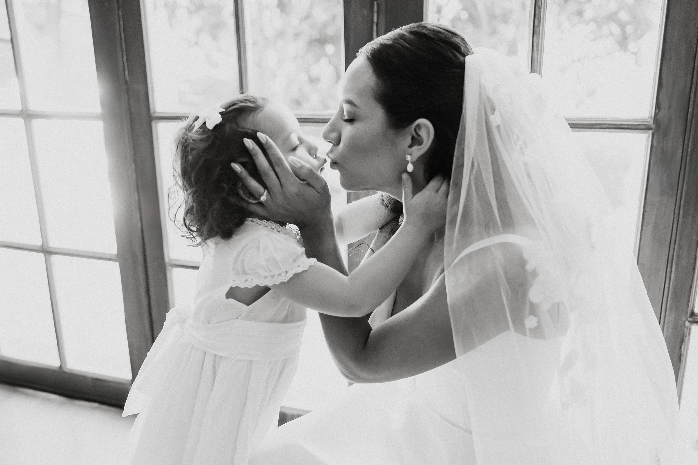 Black and white photo of a bride in a veil sharing a tender kiss with a young flower girl in a white dress.