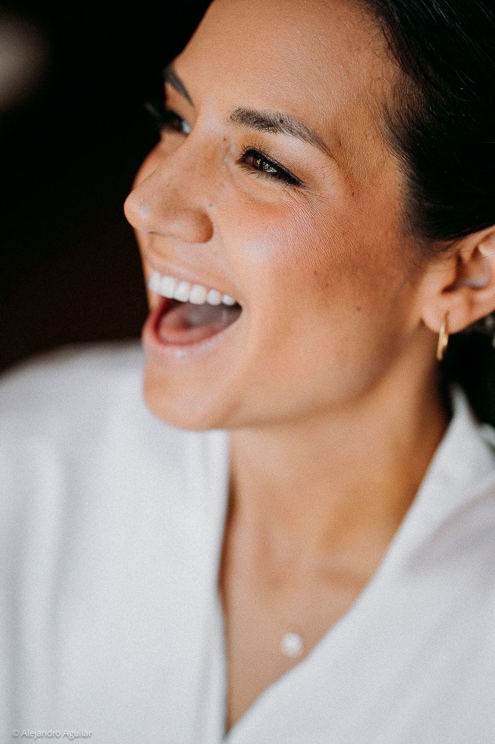 A woman laughing joyfully, wearing gold hoop earrings and a white top, with natural glowing makeup.