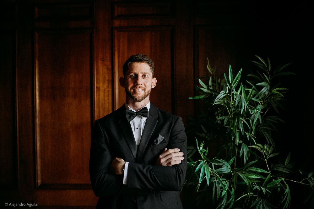 Groom in black tuxedo with bow tie stands arms crossed beside greenery against wooden wall.
