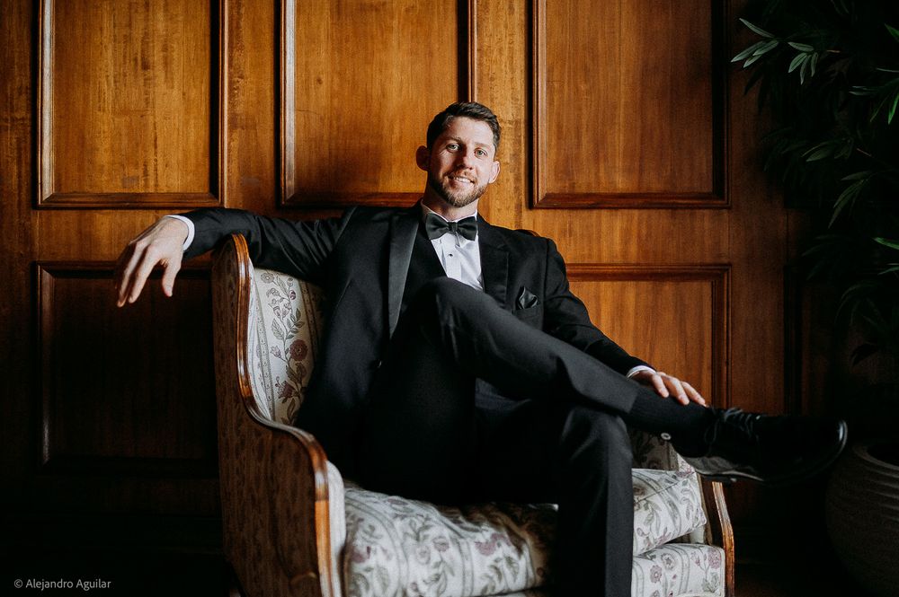 Groom in black tuxedo and bow tie sitting relaxed in ornate chair against wood-paneled wall.