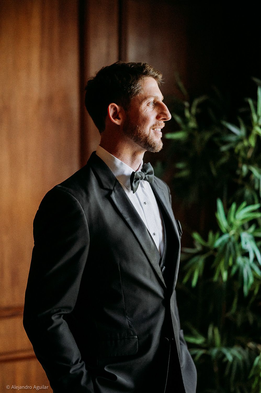Groom in black tuxedo and bow tie standing elegantly near wooden wall and green plants.