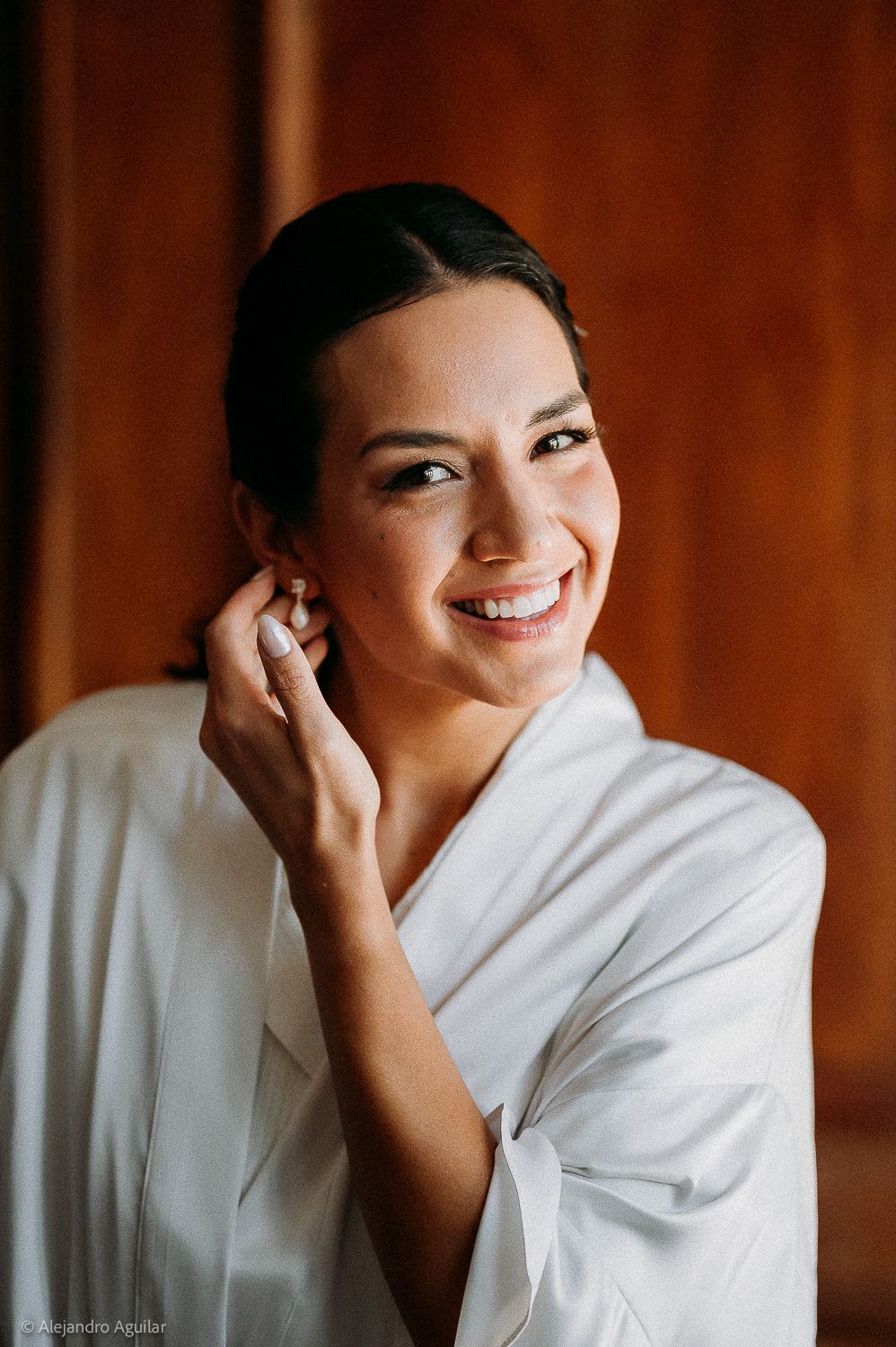 Smiling bride in white robe putting on pearl earring, with elegant updo hairstyle and natural makeup.