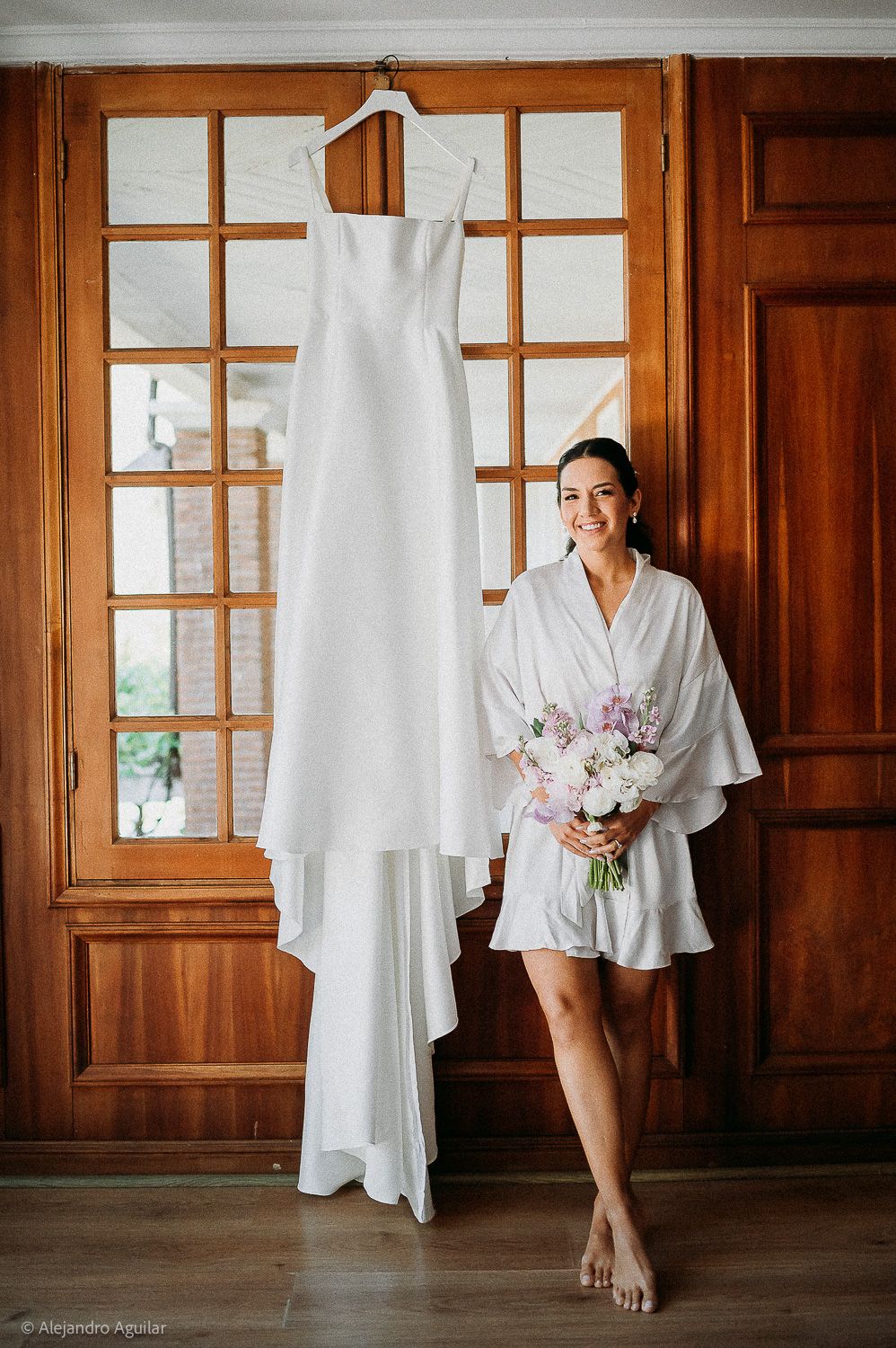 Bride in white robe holding floral bouquet stands beside elegant sleeveless wedding gown hanging on wooden door.