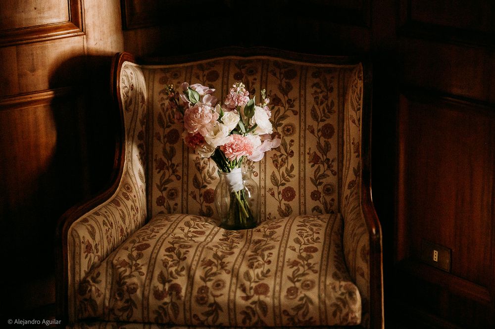 Elegant floral bouquet with pink and white flowers in a glass vase on a vintage ornate armchair in a dimly lit room.