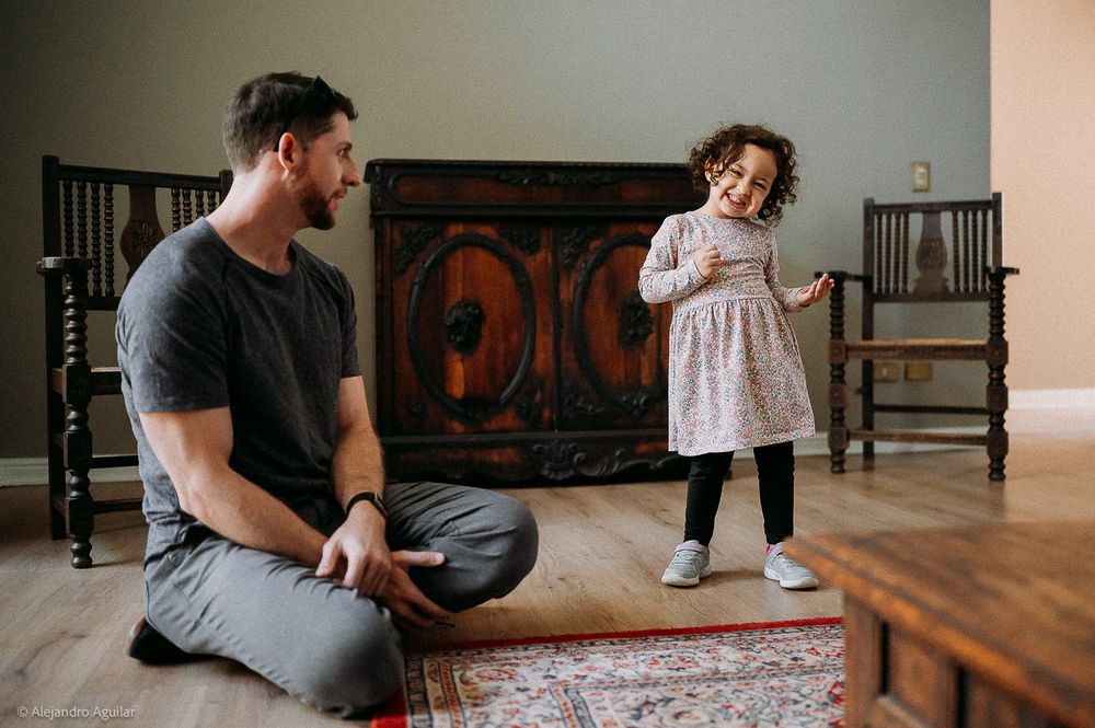 Father sitting on floor smiling at young daughter in floral dress dancing in cozy living room.