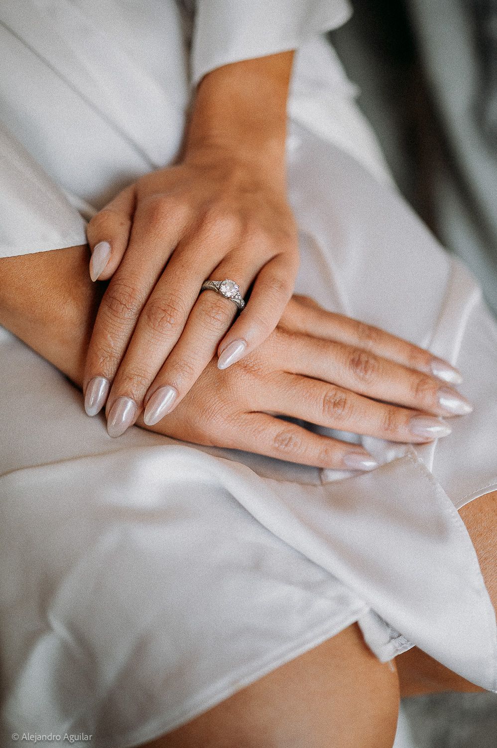Close-up of a bride's hands with nude almond nails and a diamond engagement ring resting on a white satin robe.