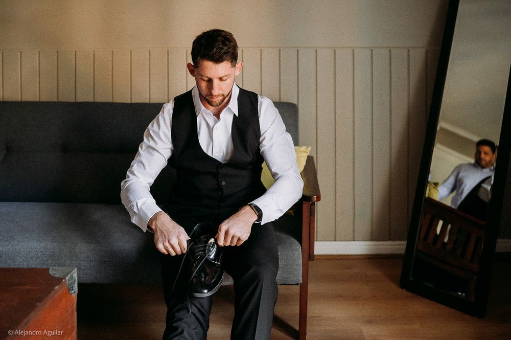 Groom in white shirt and black vest sitting on sofa putting on dress shoes before wedding ceremony.