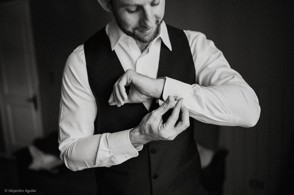 Groom in black vest fastening cufflinks on white dress shirt, black and white wedding preparation photo.